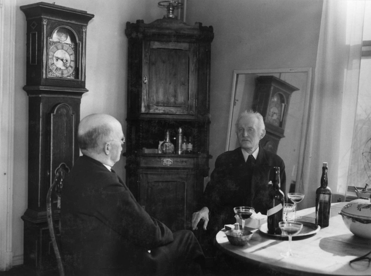 Anders Beer Wilse, Kunstneren og maleren Edvard Munch fotografert i 1937 på Ekely. Sitter ved spisebordet i samtale med fotograf Anders Beer Wilse. Flasker og tre glass til servering på bordet. Interiør med klokke og hjørneskap ved vindu.
