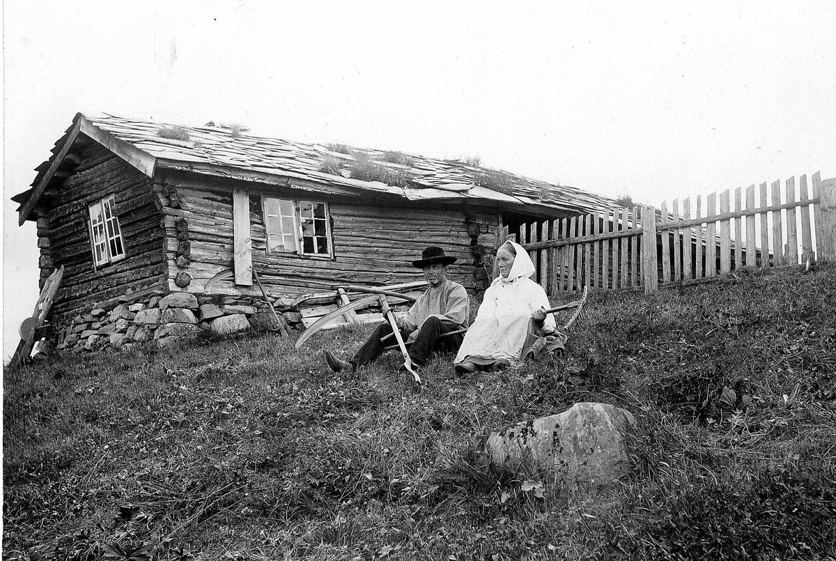 Robert Collett, Slåttefolk, antatt, sittende utenfor våningshus, ukjent sted, med ljå og rive. Slipestein står opp mot husveggen. Stakittgjerde.Huset har skifertak. Serie tatt av Robert Collett (1842-1913), amatørfotograf og professor i zoologi.