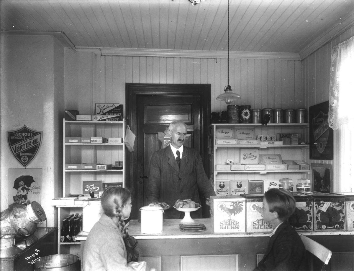 Anders Beer Wilse, Fredbo landhandleri på Høybråten i Oslo. Jente og gutt blir ekspedert av mann bak disken. Kolonialvarer i hyllene.