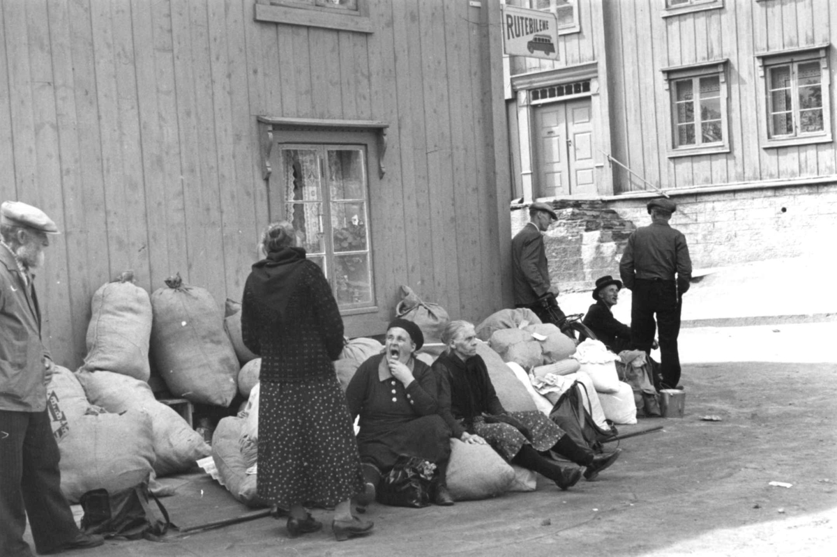 Anna Grostøl, Gatebilde med menn og kvinner som står og sitter ved fulle striesekker og annen bagasje, På bygningen henger et skilt hvor det står "Rutebilene". Røros 1938.