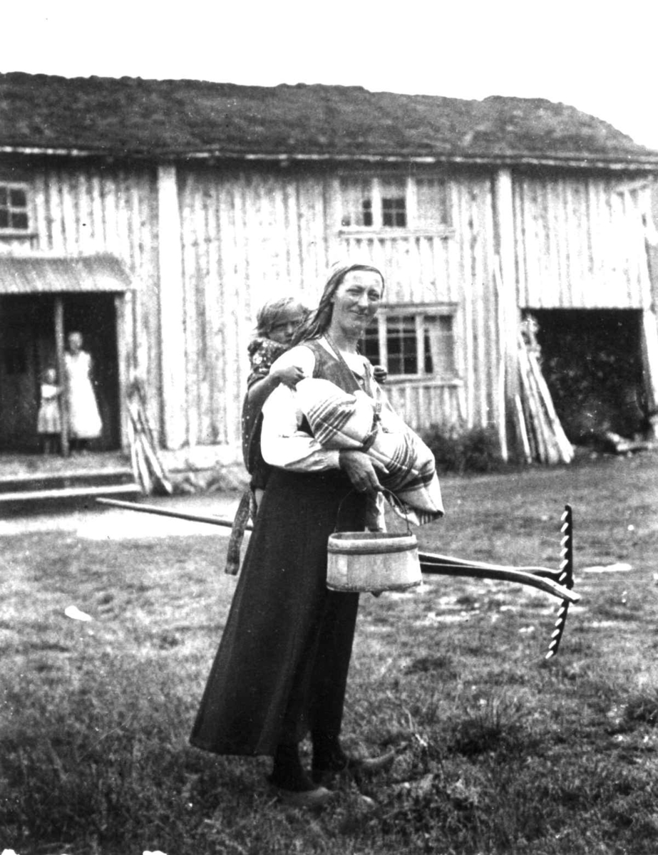 Anna Grostøl, Oline Seldal med barn i fatle, i Sirdal kalt linde, et barn på ryggen og bøtte og rive i hendene, Fidjeland, Sirdal, Vest-Agder 1936. Rekonstruksjon. Barnet hun har på ryggen er hennes niese, Tordis Harbo, f. Fidjeland (f. 1932).Et hus i bagrunnen med kvinne og barn i inngangspartiet. Oline Seldal …