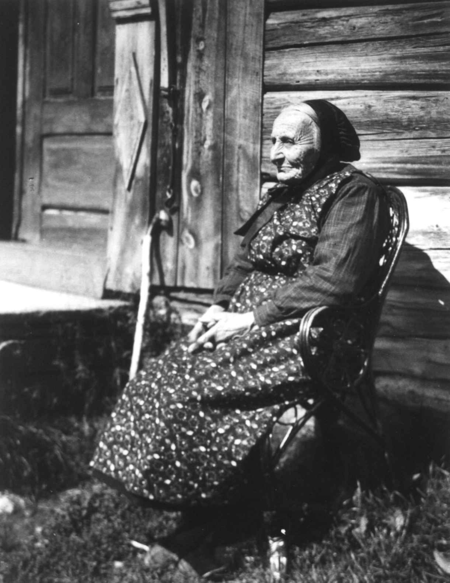 Anna Grostøl, Fru Marit Kvalsjord sitter i en kurvstol utenfor inngangspartiet til et hus. Vognill, Oppdal, Sør-Trøndelag 1934.