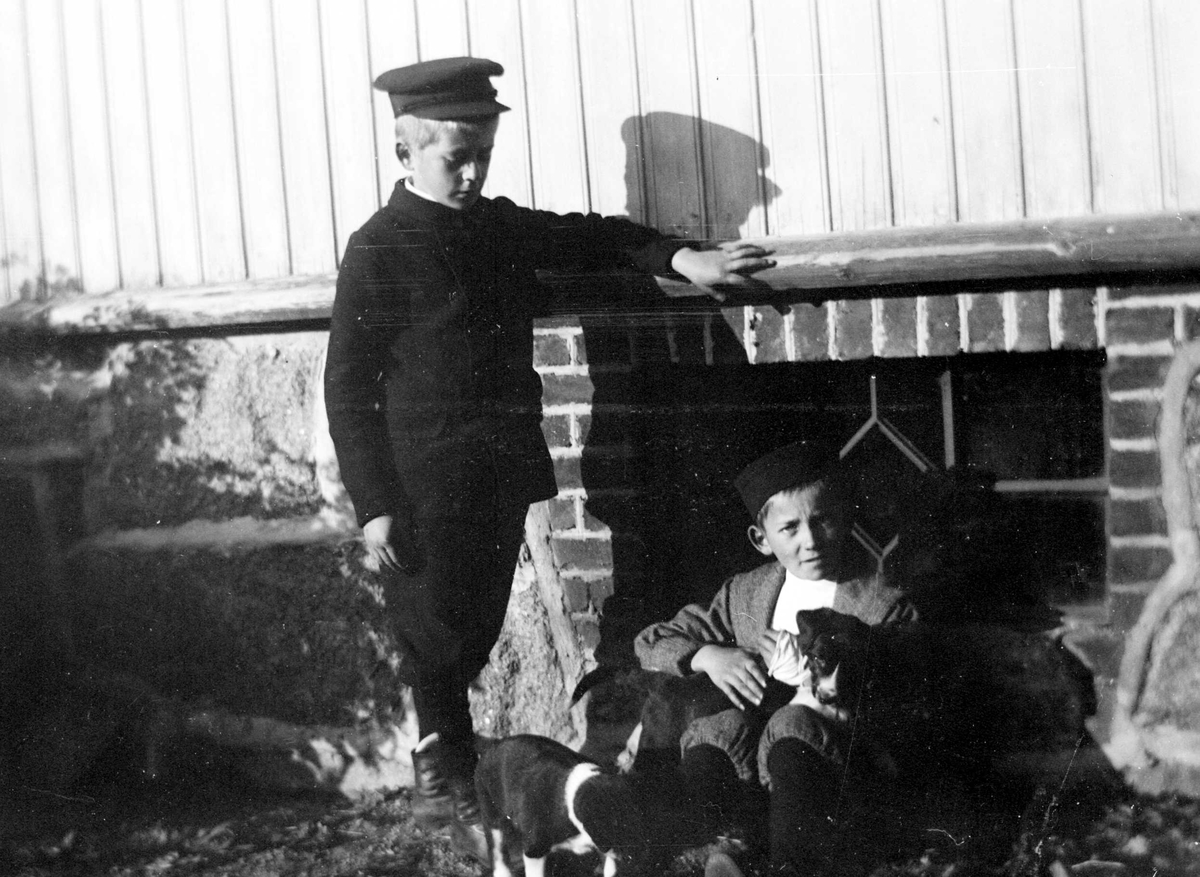 Juliane Fredrikke Langberg, Portrett av 2 gutter med skyggelue og hundevalper foran husmur med vindu med jernsprosser. Christian L. Holm t.v. er fotografens nevø, sønn av sognepresten. Gutten t.h. er sønn av klokker Busland i Hurum. Ant. på Hurum prestegård. Fra malerinne Juliane Fredrikke Langbergs (1856-1930) samling, tatt …