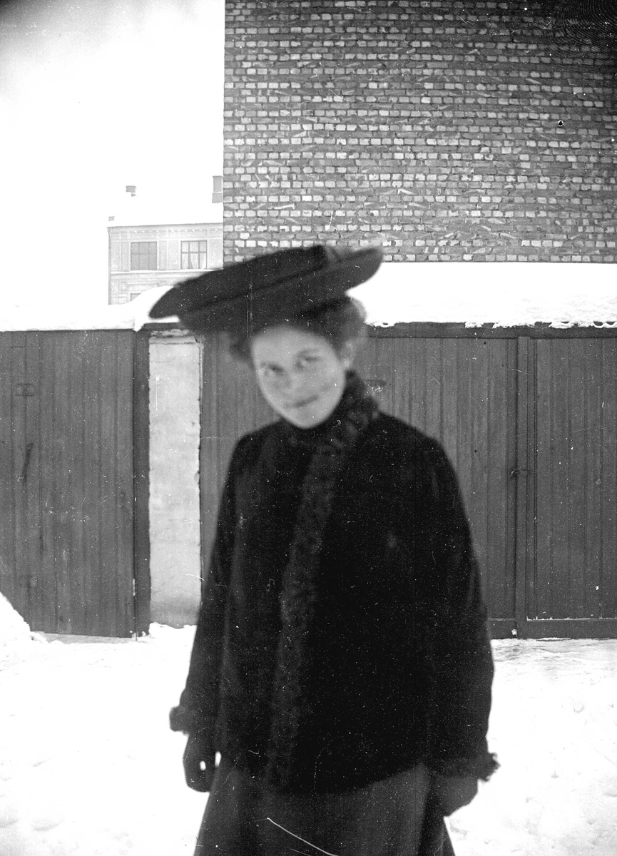 Juliane Fredrikke Langberg, Portrett av ung kvinne, Emily Holm, fotografens niese. Fra bakgård med snø i bymiljø med doer eller boder i bakgrunnen. Hun er kledd i hatt og vinterkåpe med pelskanter. Noe uskarpt bilde. Fra malerinne Juliane Fredrikke Langbergs (1856-1930) samling, tatt ca.1895-1910 med motivkrets slekt og venne…
