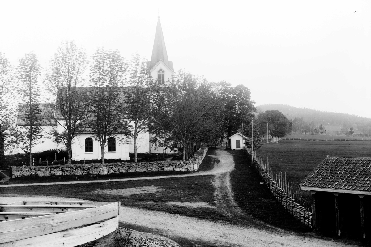 Juliane Fredrikke Langberg, Hurum kirke med omgivelser. Steinmur omkring kirken, skigard ved veien. I forgrunnen t.v. ligger muligens snøplog av tre. Fotografens svoger, Olaf A. Holm, var sokneprest i Hurum 1897-1904. Fra malerinne Juliane Fredrikke Langbergs (1856-1930) samling, tatt ca.1895-1910 med motivkrets slekt og venn…