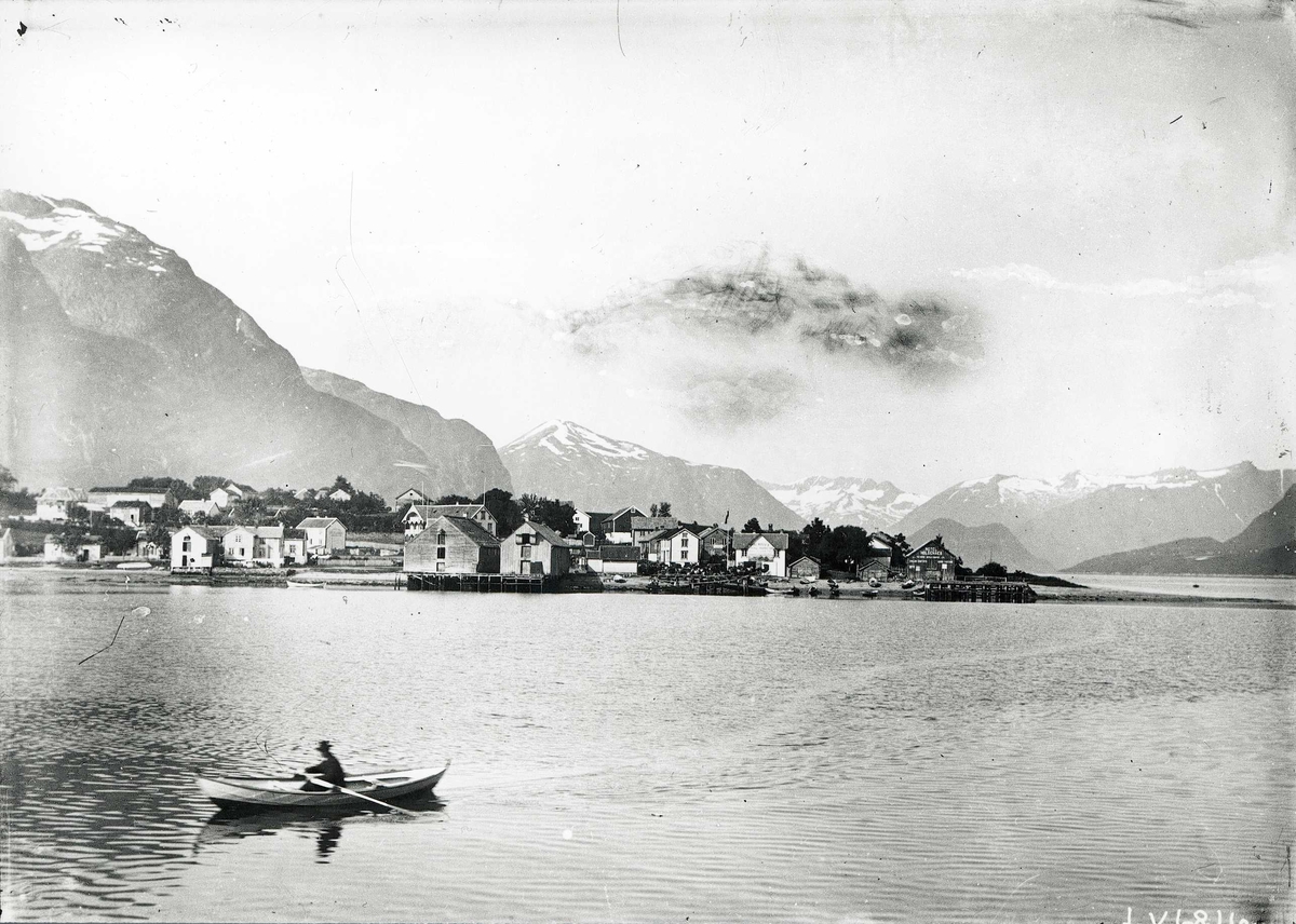Tom Kent, Kystbebyggelse fra sjøen. Boliger og sjøboder mot fjell med snø. Mann i robåt.