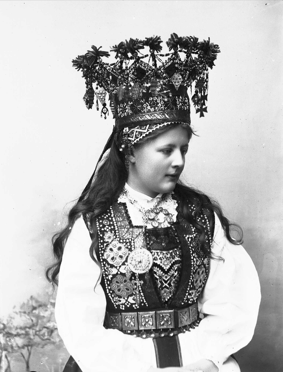 Solveig Lund, Studiofotografi av kvinne med brudedrakt og brudekrone Sittende forann malt bakgrunn med blomster/plante motiv. Hofteutsnitt. Vendt mot høyre. Ser nedover.