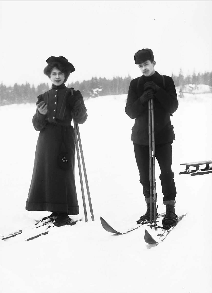 Solveig Lund, Mann og kvinne i snødekt landskap. Fotografert forfra - begge med ski på beina. I bakgrunnen står en kjelke.