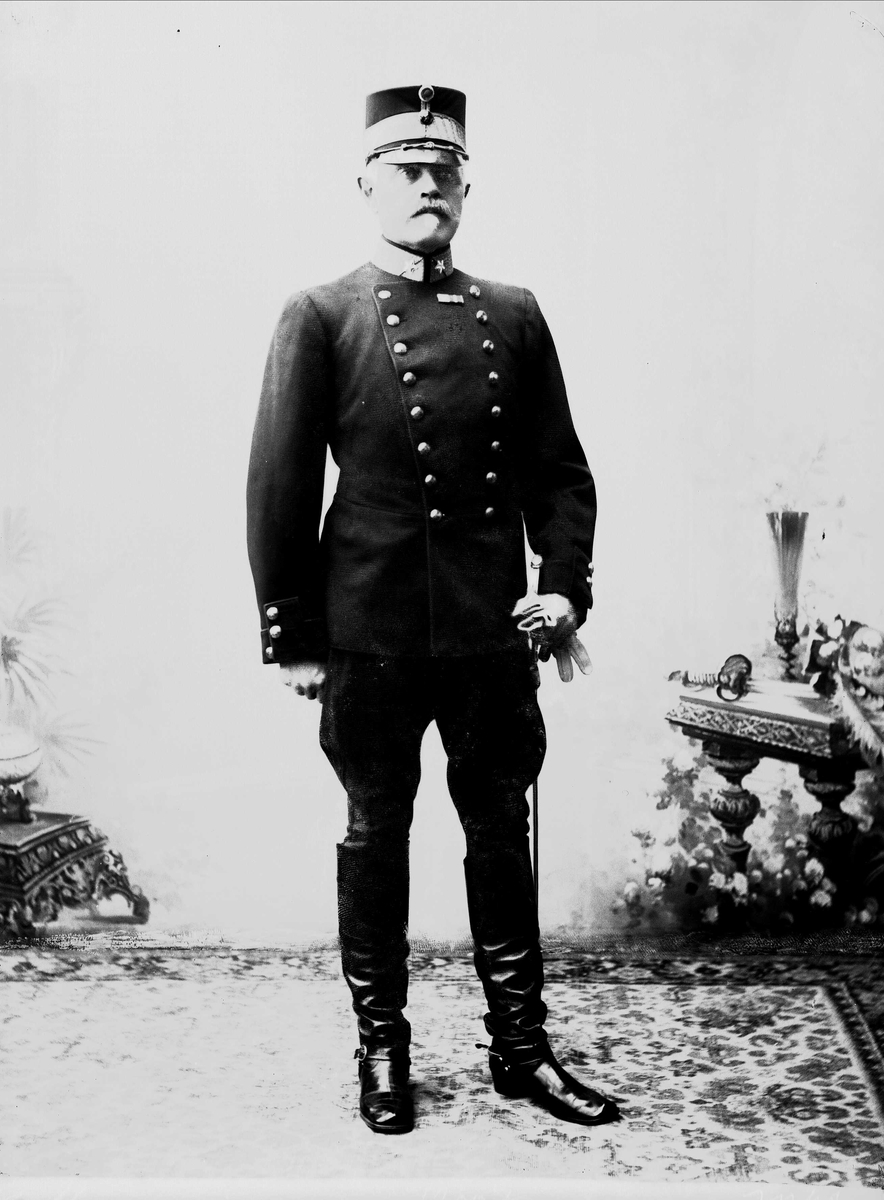 Solveig Lund, Studioportrett av eldre mann i uniform stående på teppe foran malt bakgrunn med bord, vase og blomster.