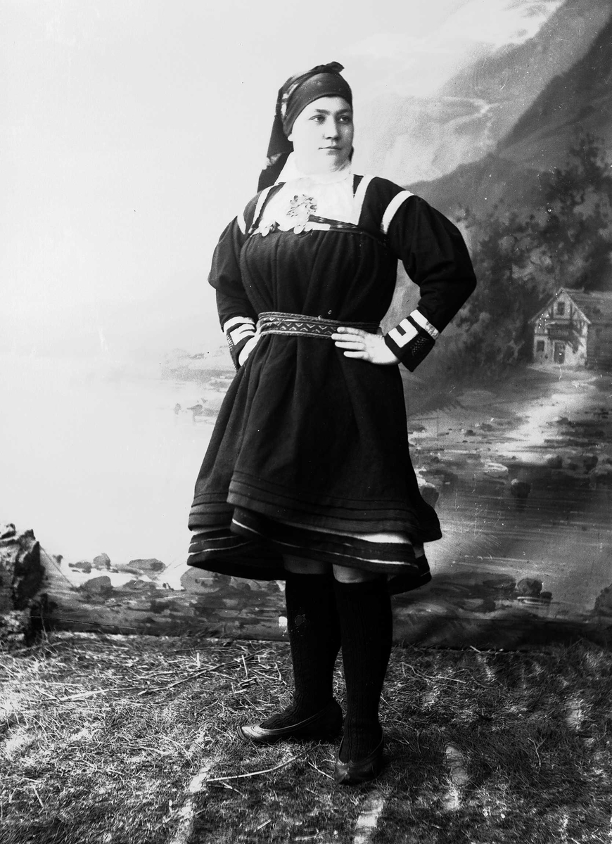 Solveig Lund, Studioportrett av kvinne i nasjonaldrakt avbildet fra siden. Hun står vendt mot venstre og ser ut mot høyre. Står på gress/jordbunn. Malt bakgrunn med fjord og fjell og et lite hus.