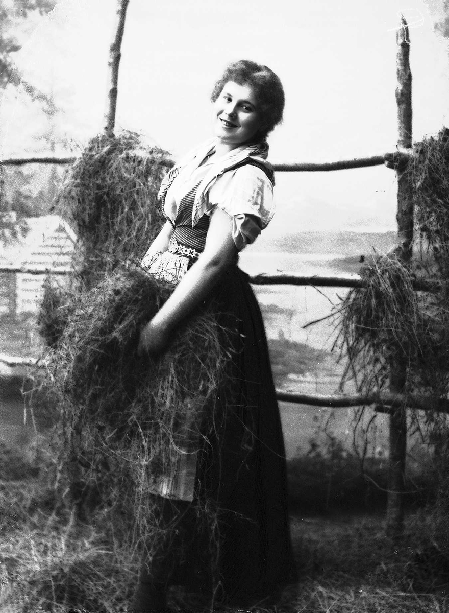 Solveig Lund, Studioportrett av kvinne i nasjonaldrakt. Hun står foran hesjegjerde og holder høy i hånden. Bakgrunnen er malt med skog, vann og hytte. På gulvet er det høy/gress.