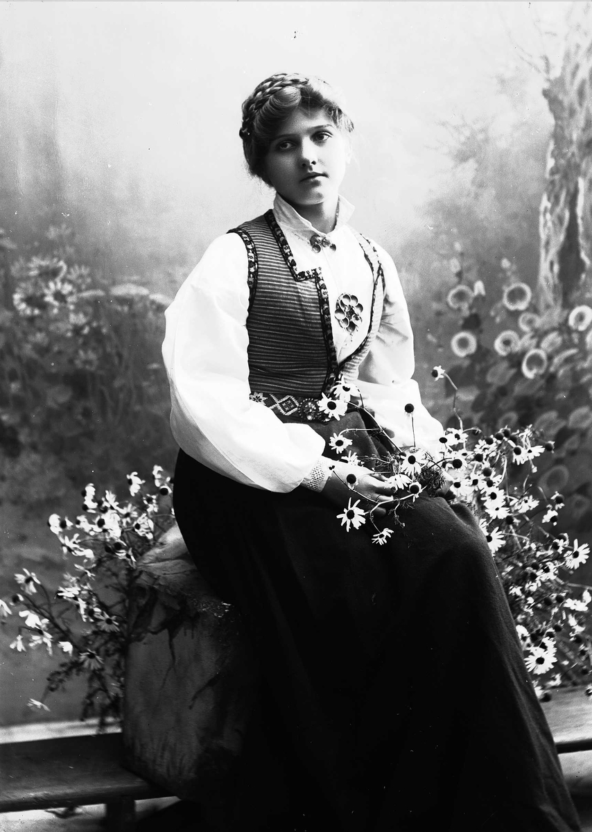 Solveig Lund, Studioportrett av kvinne i drakt sittende på modell av stein. Hun holder blomster i hendene.Bakgrunnen er malt med trær og blomster.