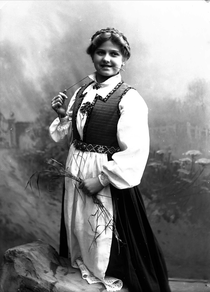 Solveig Lund, Studioportrett av kvinne i drakt. Holder strå i hendene som hun tygger på. Bakgrunnen er malt med blomster.