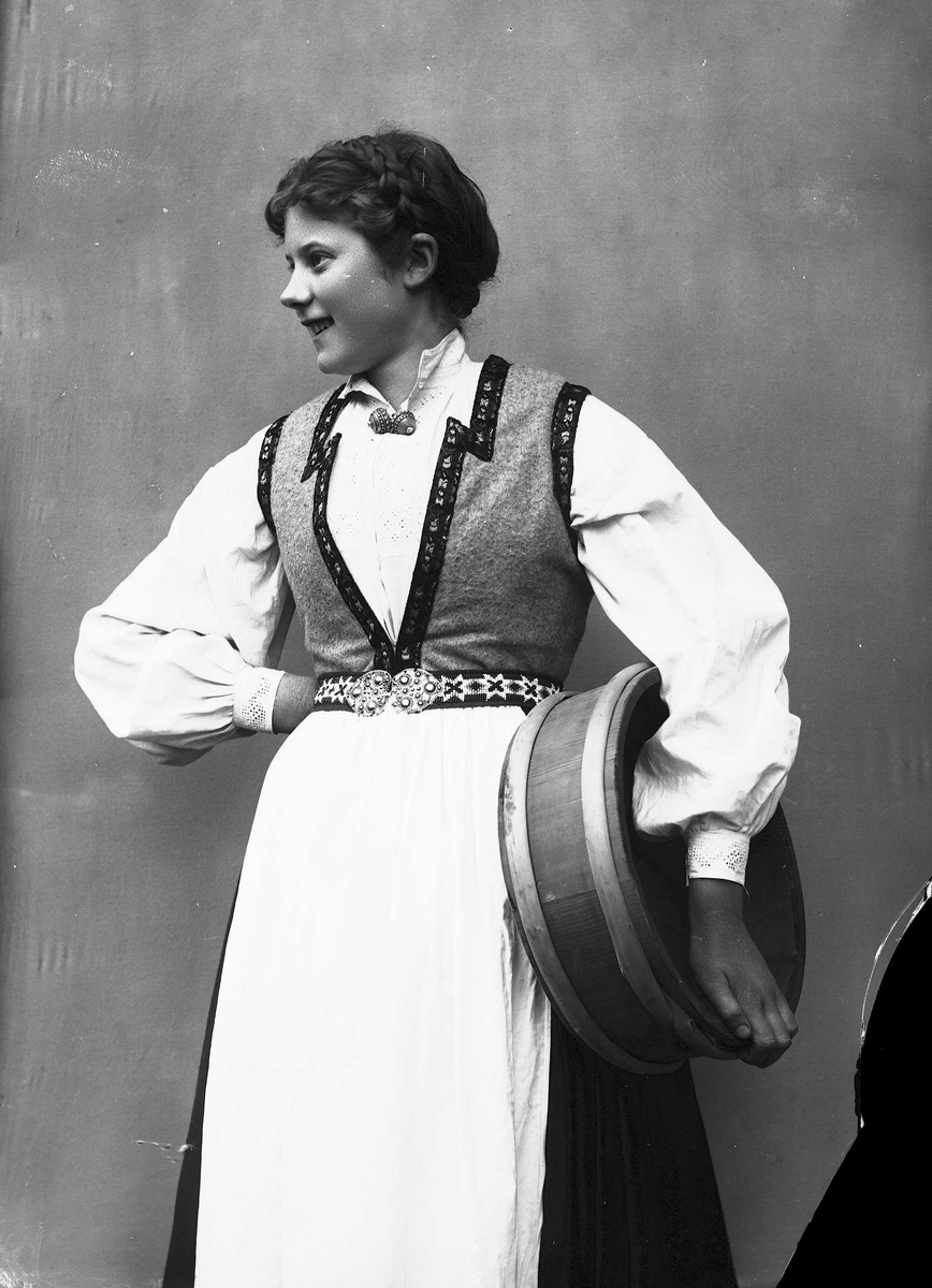 Solveig Lund, 3/4 portrett av kvinne i drakt, tatt i fotografens studio. Hun holder en melkekolle under den venstre armen og den høyre er satt i hoften. Hun ser ut mot venstre i bildet. Nøytral bakgrunn.