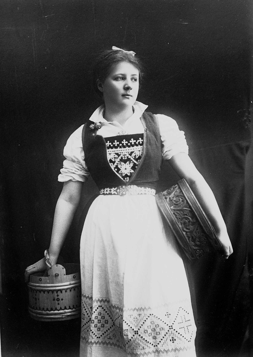 Solveig Lund, 3/4 portrett av kvinne i drakt med opprullede armer, tatt i fotografens studio. Hun holder en eske under den venstre armen og i den høyre en bøtte. Hun ser ut mot høyre i bildet. Nøytral bakgrunn.