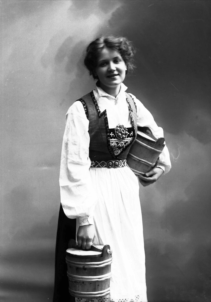 Solveig Lund, 3/4 portrett av kvinne i drakt, tatt i fotografens studio. Hun holder en tine i høyre hånd og en under venstre arm. Nøytral bakgrunn.
