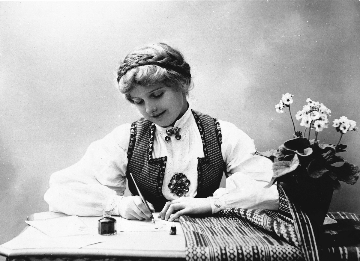 Solveig Lund, Studioportrett av kvinne i drakt med oppsatt hår. Hun sitter ved et bord og skriver et brev med penn og blekk. På bordet er det duk og blomsterpotte.