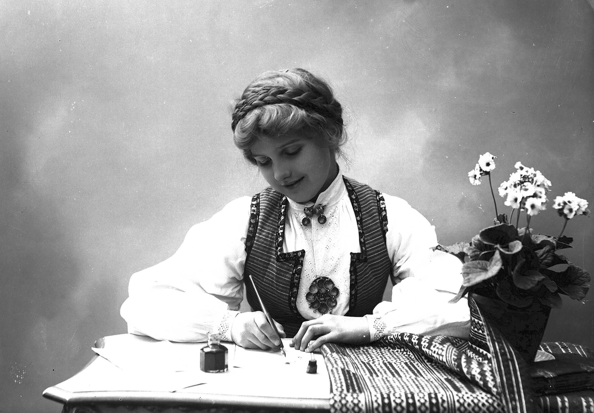 Solveig Lund, Studioportrett av kvinne i drakt med oppsatt hår. Hun sitter ved et bord og skriver et brev med penn og blekk. På bordet er det duk og blomsterpotte.