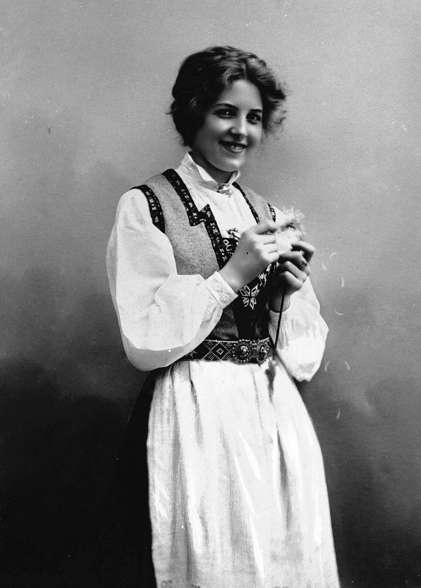 Solveig Lund, 3/4 dels studioportrett av kvinne i drakt og oppsatt hår. I hendene holder hun en blomst. Bakgrunnen er malt med himmel og skyer.
