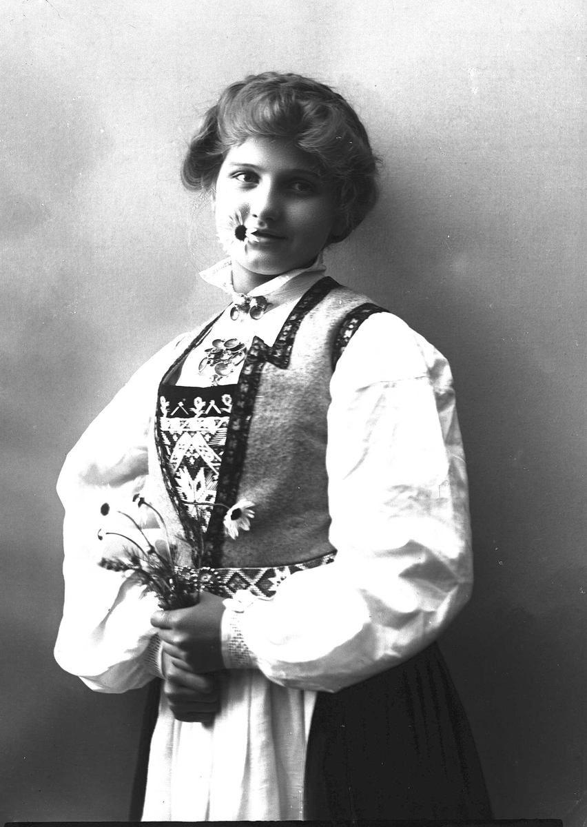 Solveig Lund, 1/2 portrett av kvinne i drakt med oppsatt hår, fotografert i studio. I munnen holder hun en prestekrage og i hendene en bukett av dem. Kvinnen ser ut av fotografiet. Nøytral bakgrunn.