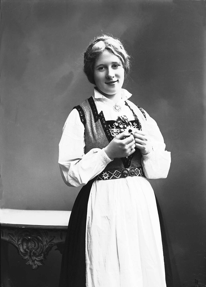 Solveig Lund, 3/4 del studioportrett av kvinne i drakt med oppsatt hår. Kvinnen lener seg mot et bord. I hendene holder hun en prestekrage. Kvinnen ser ut av fotografiet. Nøytral bakgrunn.