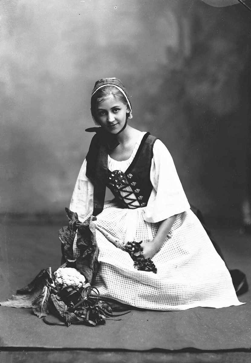 Solveig Lund, Studioportrett av kvinne i drakt og skaut. Hun sitter på knærne på et teppe, foran en haug med grønnsaker. Avfotografert foran en nøytral bakgrunn.