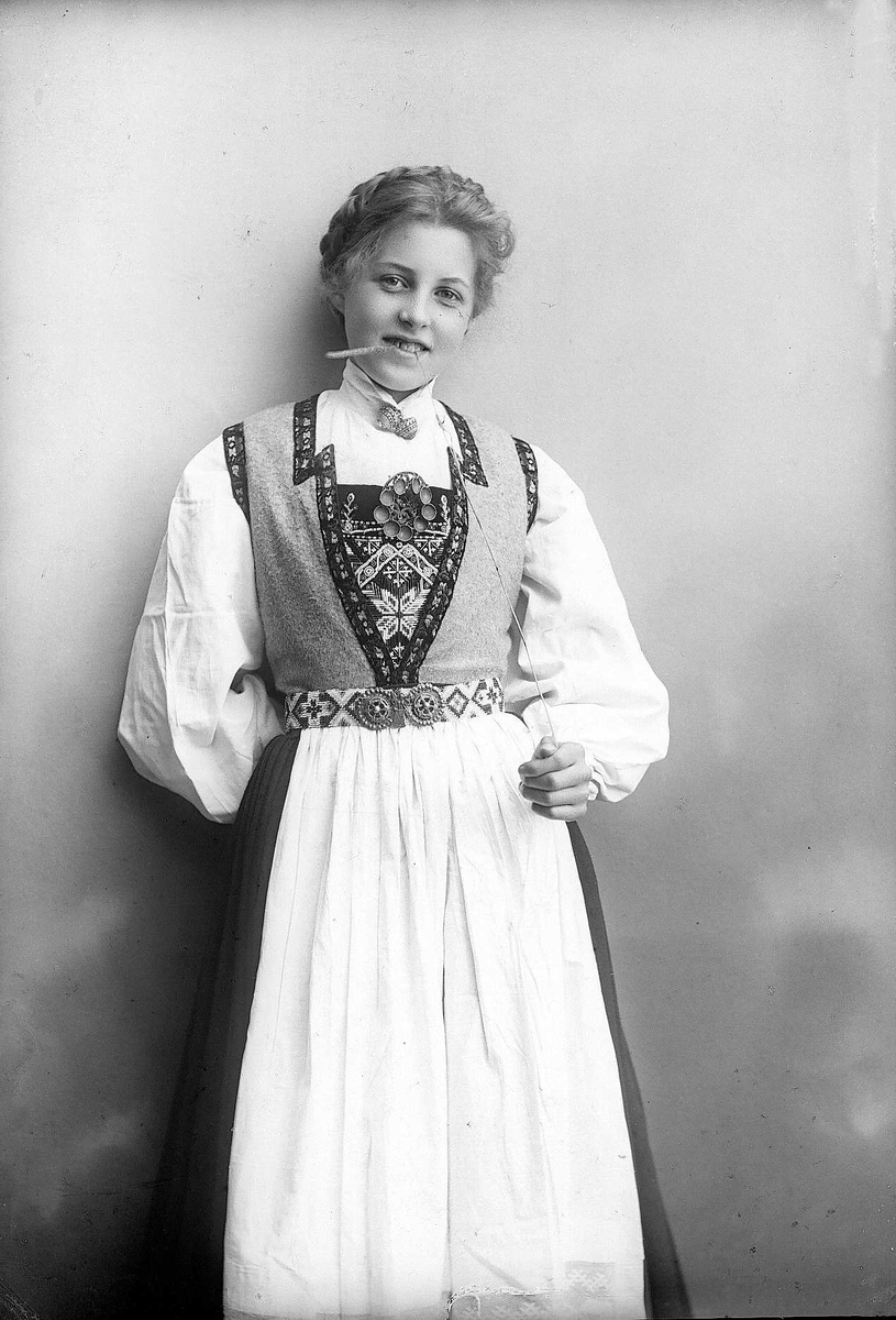 Solveig Lund, 3/4 dels studioportrett av kvinne i drakt, med oppsatt hår. Hun biter på et strå hun holder i venstre hånd. Hun er avfotografert mot en nøytral bakgrunn.