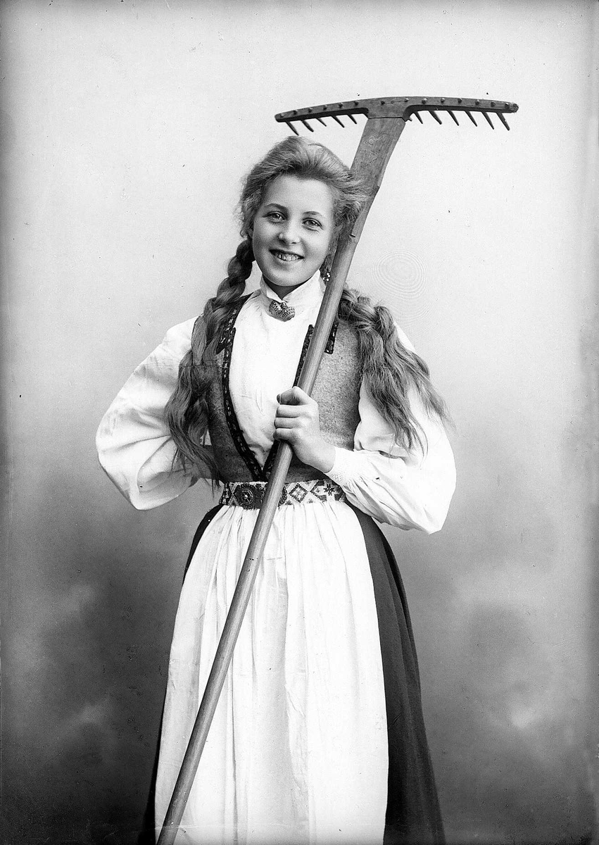 Solveig Lund, 3/4 dels studioportrett av kvinne i drakt, med flettet hår. Hun holder en rive i venstre arm, mens den høyre hviler mot hoften. Bakgrunnen er nøytral.