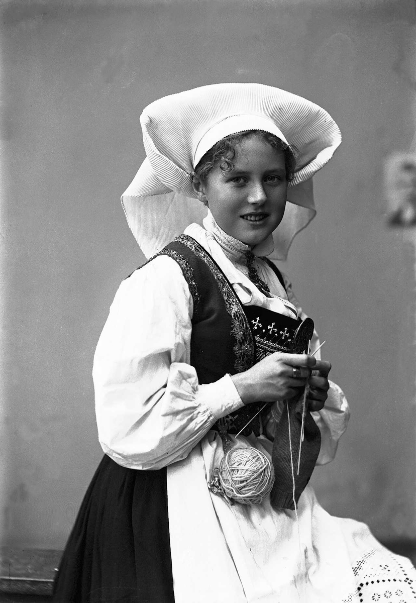 Solveig Lund, 1/2 dels studioportrett av kvinne i drakt og skaut, som sitter på en benk og strikker. Bakgrunnen er nøytral.