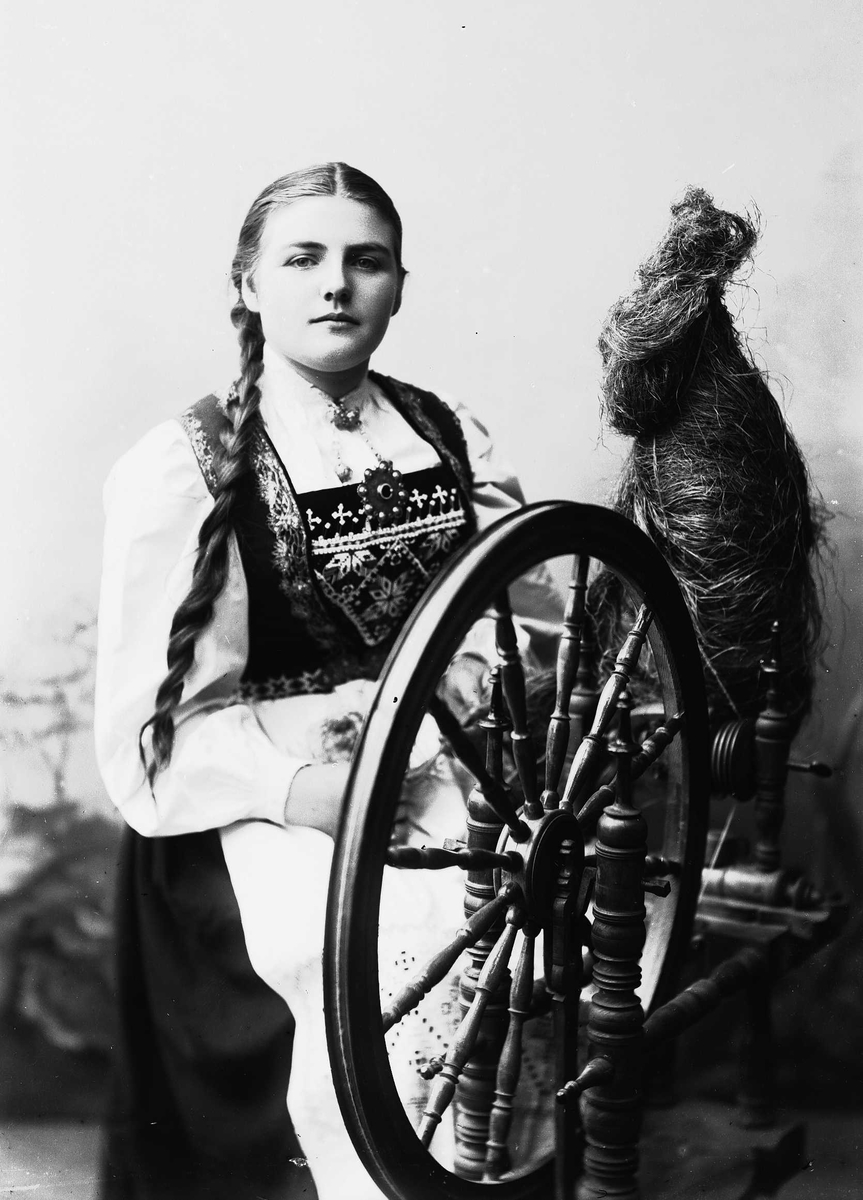 Solveig Lund, Studioportrett av kvinne i drakt og flettet hår, som sitter ved en rokk. Bakgrunnen er nøytral.