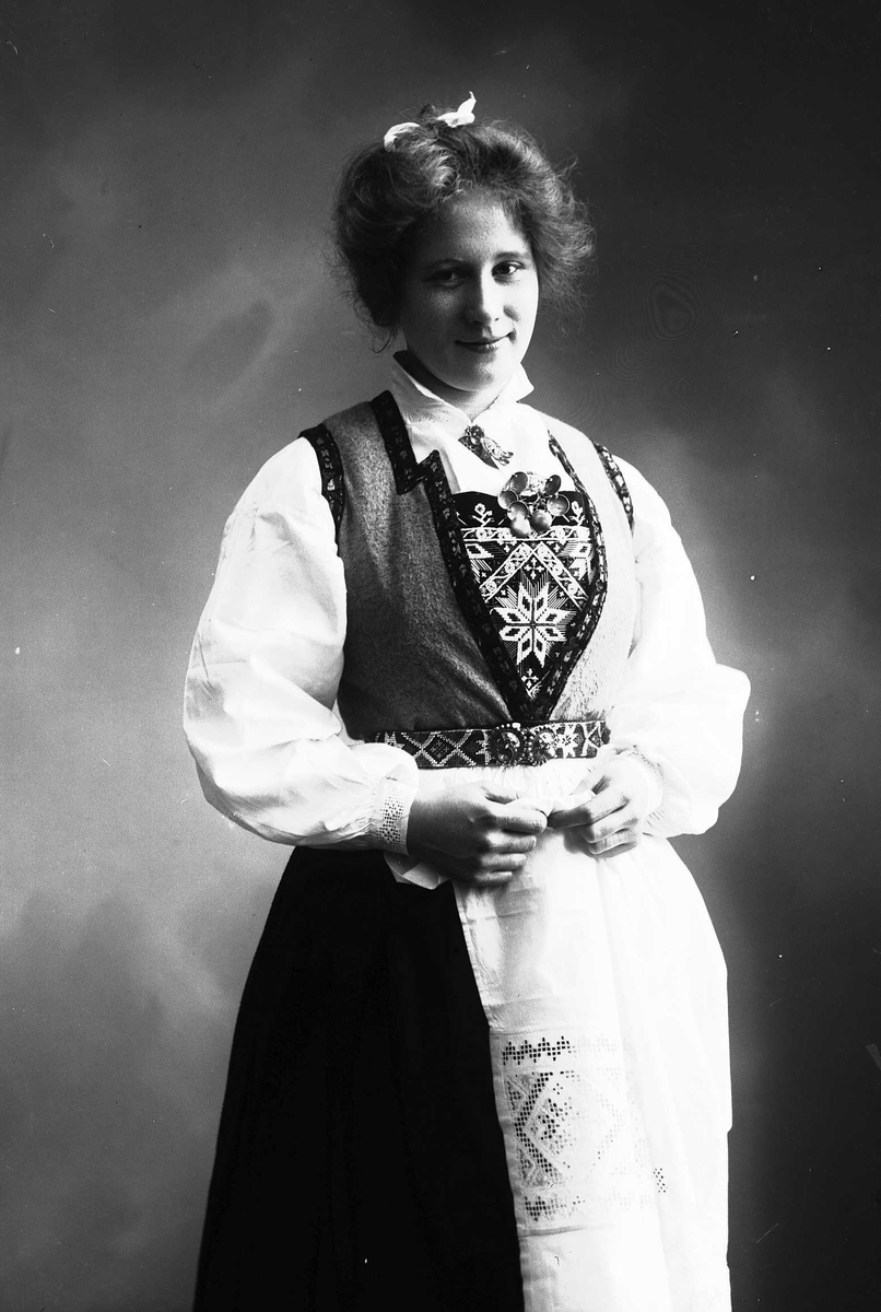 Solveig Lund, Studioportrett av kvinne i drakt med sløyfe i håret. Hun holder forkle sitt i hendene.