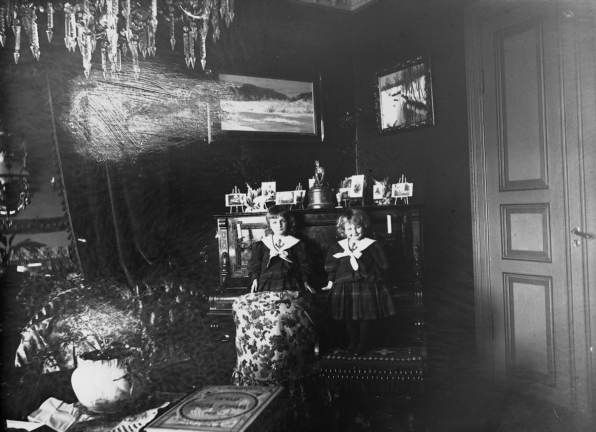 Axel Quinsgaard Wiborg, Karen og Gudrun Q. Wiborg sitter på piano i stue, Meltzers gate 9, Oslo, 1899. Fotografier i ramme plassert på pianoet.