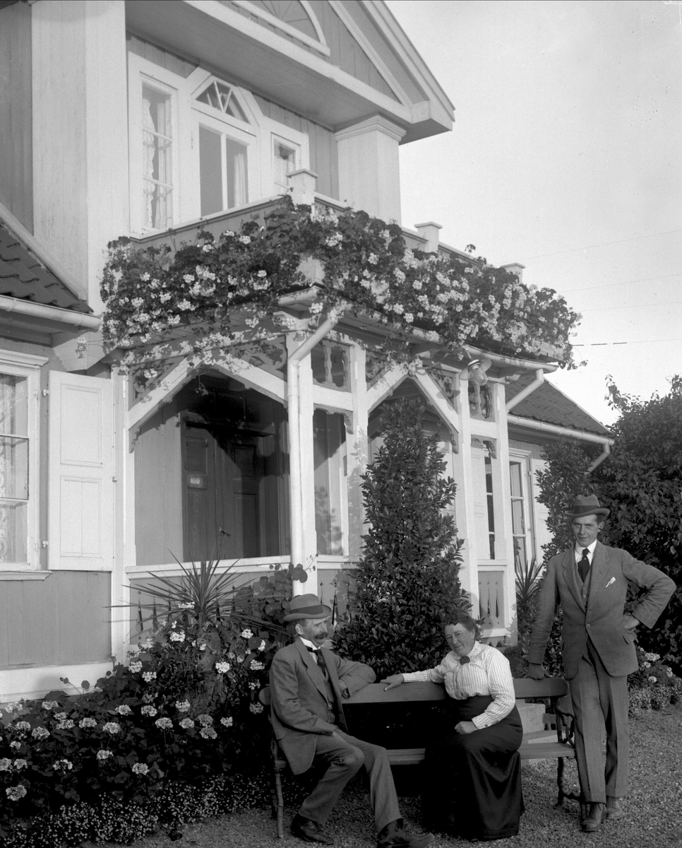 Gustav Borgen, Gruppe, to menn og en kvinne ved en benk foran inngangen på en villa. Blomster rundt og på verandaen.