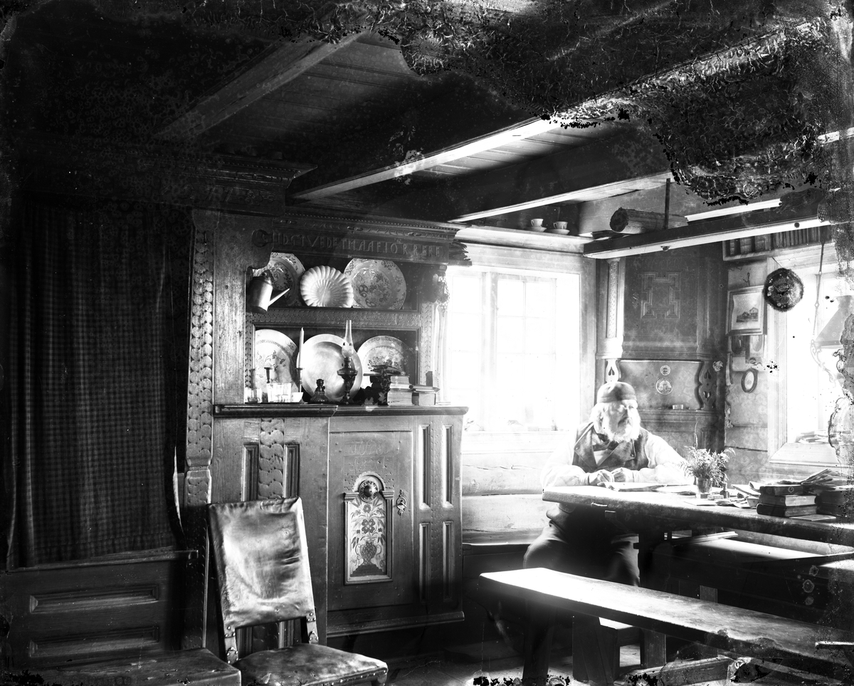 Gustav Borgen, Tømmerstue, stueinteriør med langbord, hjørneskap og framskap. Eldre mann ved enden av bordet.
