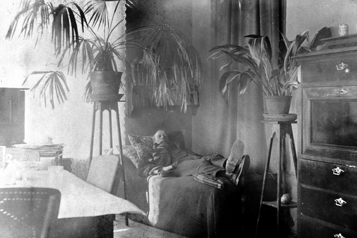 Anno Domkirkeodden, INTERIØR DAGLIGSTUE, MANN M/PIPE HVILER PÅ divan/SOFA, PLANTER PÅ PIDESTALLER, KOMODE, STRANDGATA 15 "Divanen ble et svært populært møbel fra slutten av 1800-tallet. De var både del av stueinteriører og soveromsmøbler" Se årboka for Hedmarksmuseet/Hamar Historielag "Fra Kaupang og Bygd 2010" Artikk…