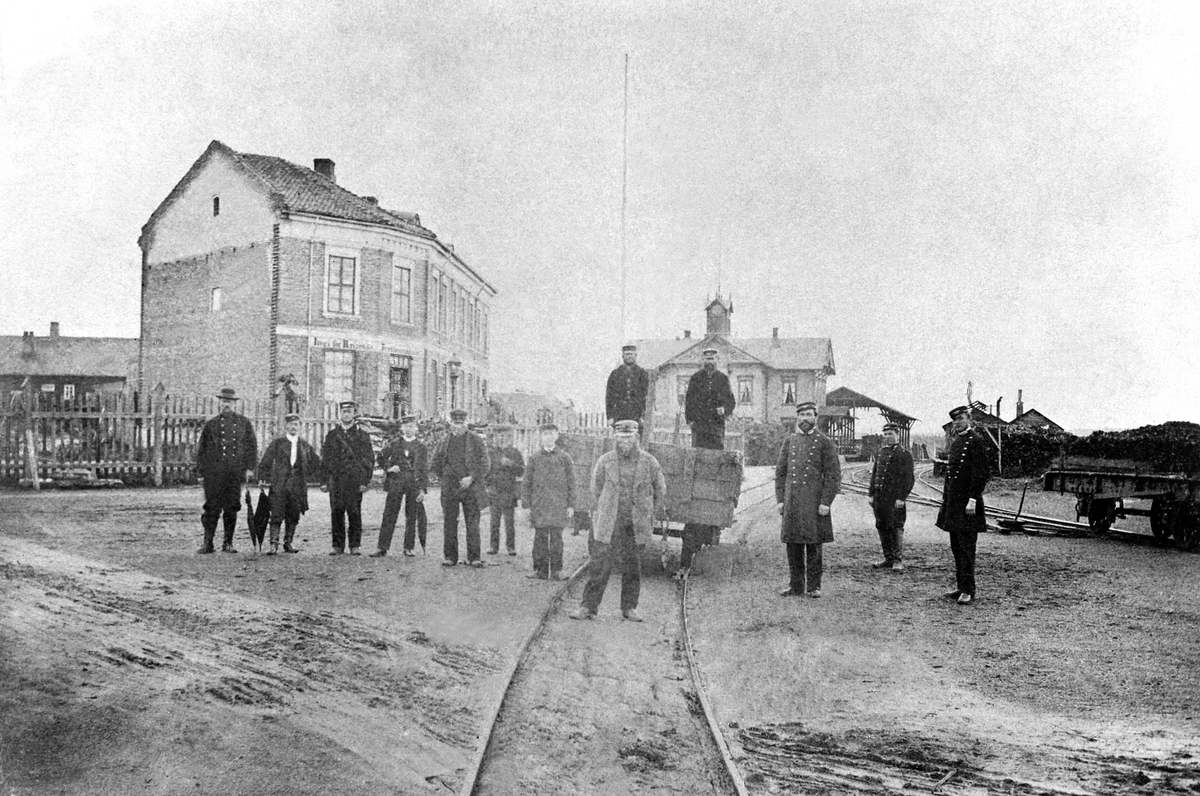Anno Domkirkeodden, Eksteriør Hamar Stasjonsbygning nr. 1 fra 1862, Kluges Hotell til venstre kalt "Jernbanehotellet", gruppe 13 personer. Banemester Ralph King står fremst midt i bildet, stasjonsmester Chr. Lund og kontorist Hans Eriksen står til høyre.