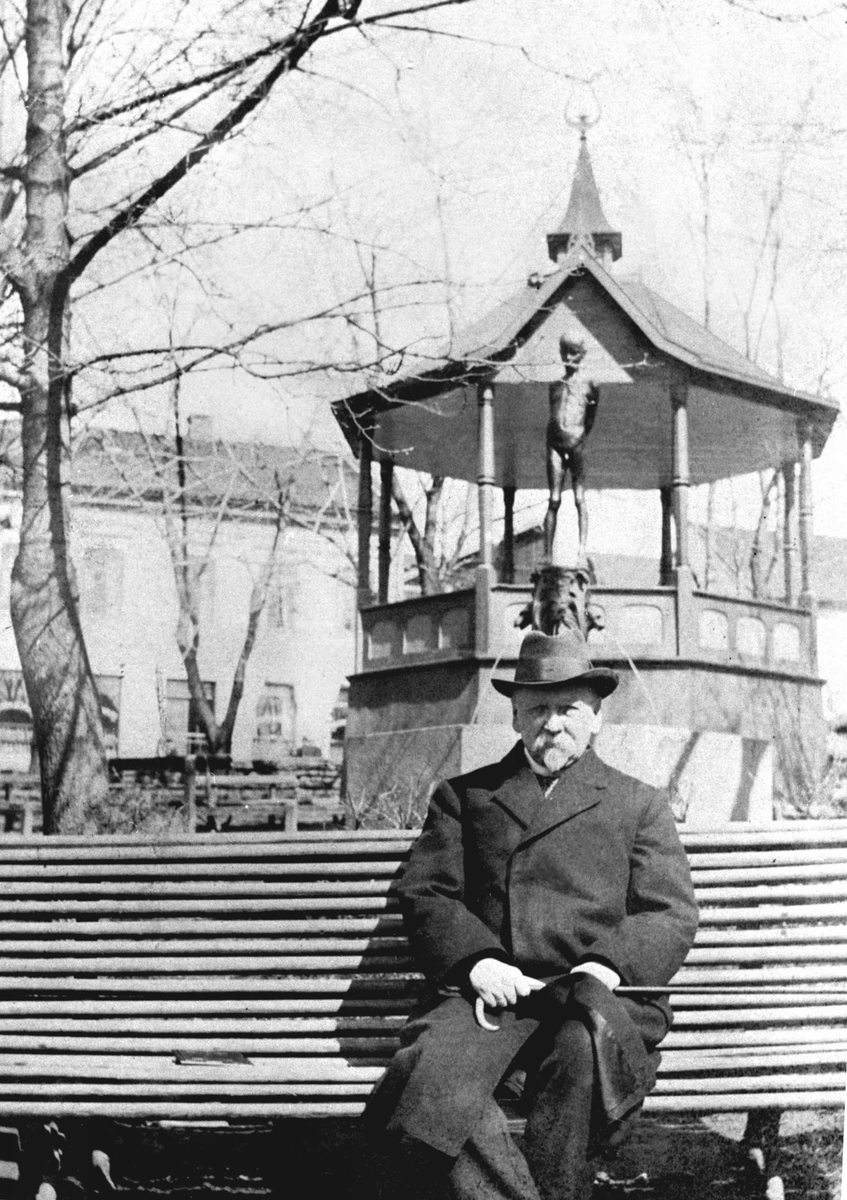 Anno Domkirkeodden, AXEL DANIEL HALVORSEN MED HATT FRAKK OG SPASERSTOKK PÅ BENK FORAN BRONSESTATUEN AV GUSTAV VIGELANDS HAMARGUTTEN PÅ TRIANGELEN OG MUSIKKPAVILJONGEN