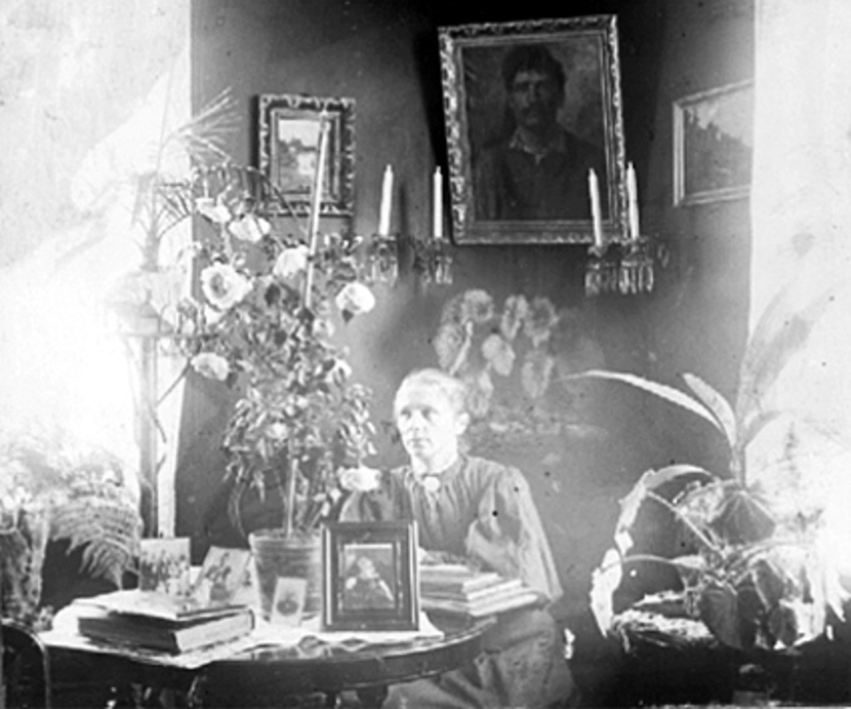Anno Domkirkeodden, INTERIØR STUE, FRU PETERSEN VED RUNDT BORD, PÅ BORDET FOTOGRAFIER, FOTOALBUM, BØKER OG EN STOR POTTEROSE, LYSESTAKER PÅ VEGGEN