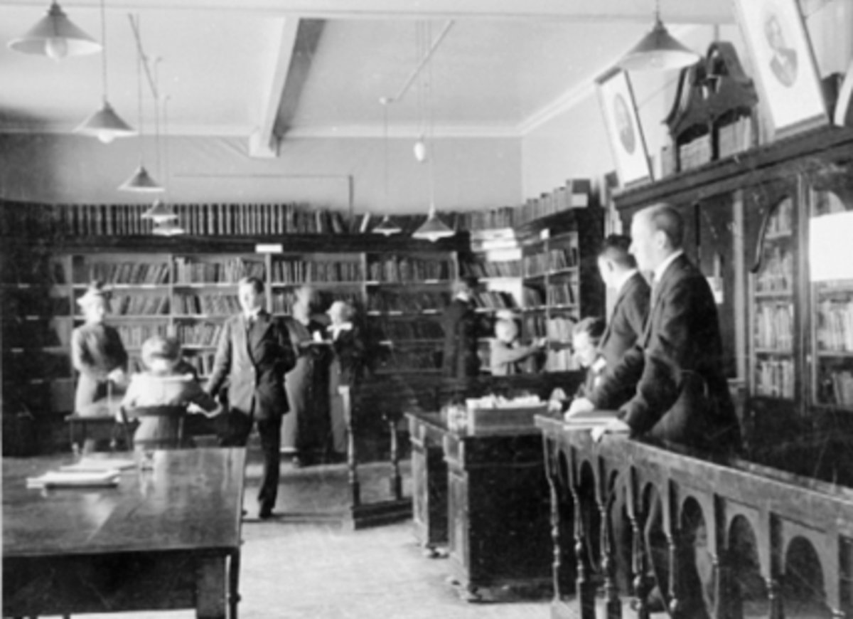 Anno Domkirkeodden, Interiør, Hamar Folkebibliotek, folk, skrivebord. Sittende v/bordet til v. Rikka Bjølgerud, stående Asbjørn Hagen Bergseng. Sittende v/utlånet Jens Jenssen. Stående Alf Melhus, Håkon Rud. Biblioteket: Brannvakta/Midtbyen 1888-1890, Håndverkern Parkgt. 1909-1925, Samlaget Torggt. 63 1925-1943 brann,…