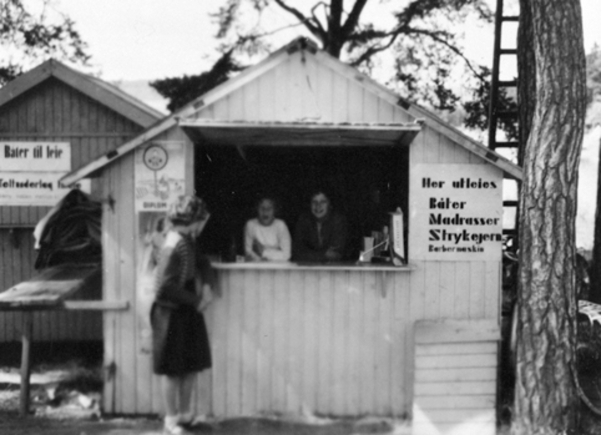 Anno Domkirkeodden, HEDMARKSMUSEET, DOMKIRKEODDEN, ROSENLUNDVIKA. KIOSK PÅ CAMPINGPLASSEN. T. V. TO UKJENTE SØATRE FRA SANDEFJORD. BEGGE PÅ BESØK TIL ONKELEN SOM DREV KIOSKEN, T. H. BERIT MOEN, PLAKAT "HER UTLEIES BÅTER, MADRASSER, STRYKEJERN, BARBERMASKIN" 1951 ELLER 1952