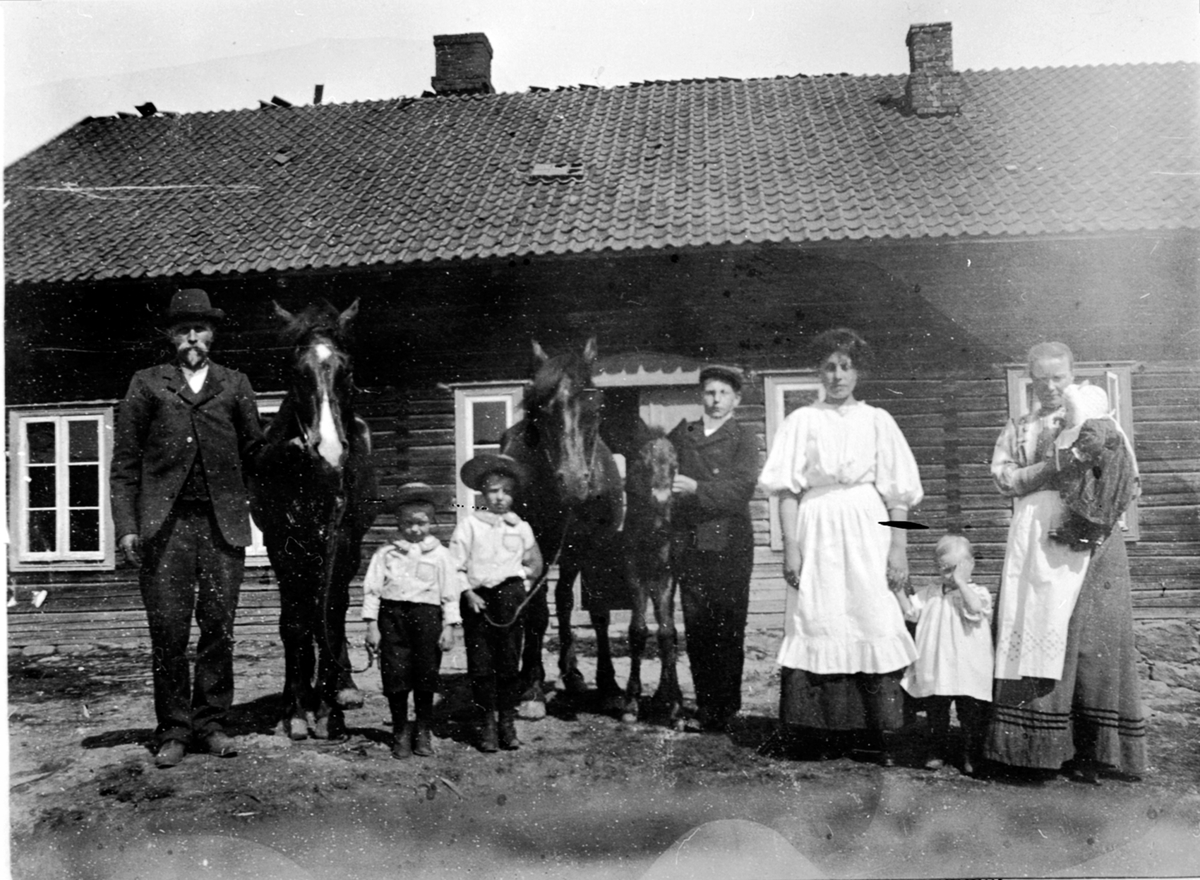 Anno Domkirkeodden, Gruppe mennesker oppstillt utenfor hus. Syverud, Ringsaker. Fra venstre er Lars Fossum f.1875, Leonard Fossum f.1906, Hans Østbye f.1905, Johan Melby f.1896, ukjent, Karen Fossum f.1909 , Gunda Fossum f.1874 med Dortha på armen. Hest med føll.