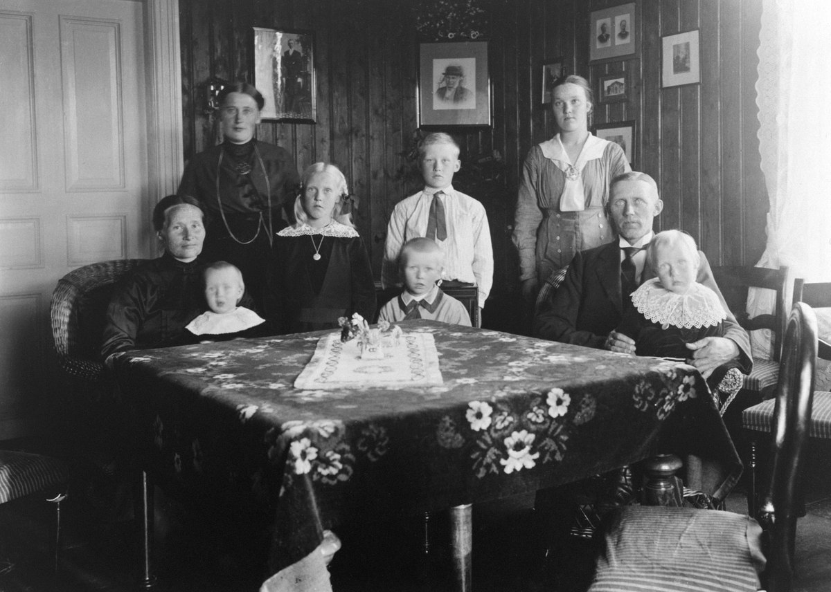Anno Domkirkeodden, Interiør, stue på Leine østre, Ringsaker. Familien Jevne. Foran er barna Håkon, Iver, Sigrid. Andre rekke fra venstre er Inga. Bak er Magnhild Bårdseth, Agnes, Erik. Bak Ester Faråsen, Ole Jevne.