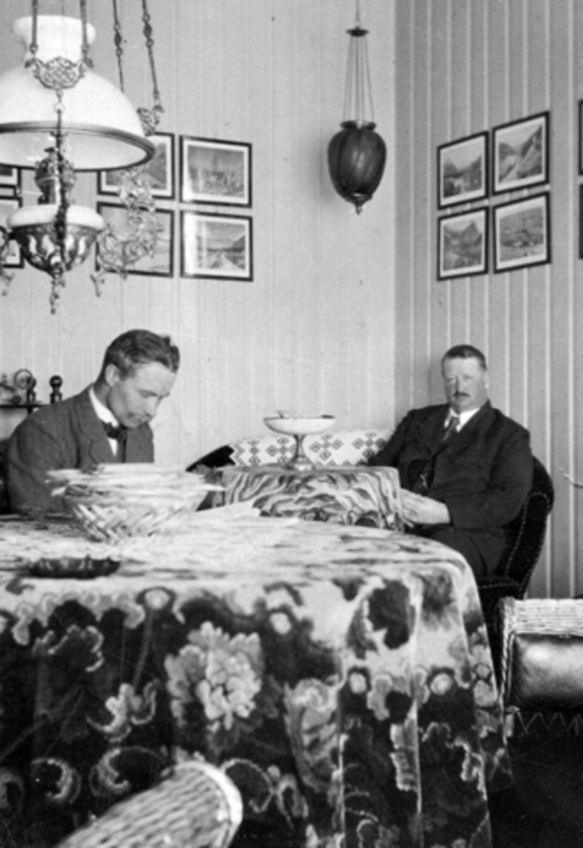 Anno Domkirkeodden, 2 menn i oppholdsrom på Toftes Gave, Helgøya. Interiør med kurvstoler, bord, parafinlampe, måneskinnslampe. Bilde på veggen.