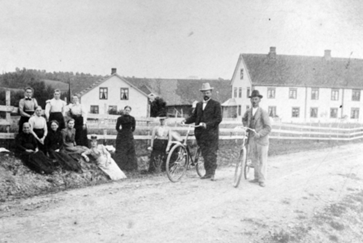 Anno Domkirkeodden, "Chauseen" på Stavsjø, Hedmark. To menn med sykkel. Fra venstre er Even Hovie (1877-1932), Mons Hjelt (1863-1959). I vegkanten fra venstre er Syverine Hovie (1881-1965), Anna Tveter (1892-1969), Karoline (1878-1960) og Anna Thorud (1872-1953). Resten er ukjente. Husene på Hjelt i bakgrunnen.