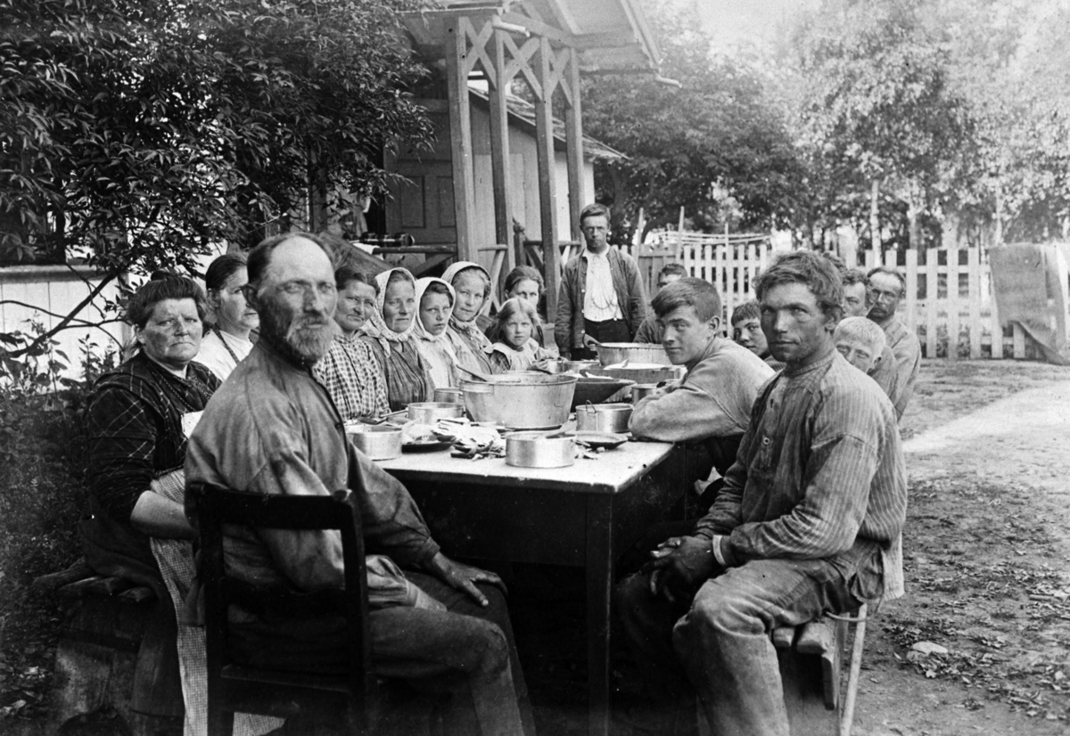 Normann Helger, Store Ree ca. 1915, måltid ute i det fri, viser gardens arbeidsstokk, gardsarbidere, tjenestefolk, boller, kjeler. Noen kjente navn, ved bordenden Einar Karlsen som bodde både i Kommat og Blæsarud og Amund Eriksen Balberg, lent innover bordet fjøsmester Hans Storsveen fra Espa, mannen som står ved …
