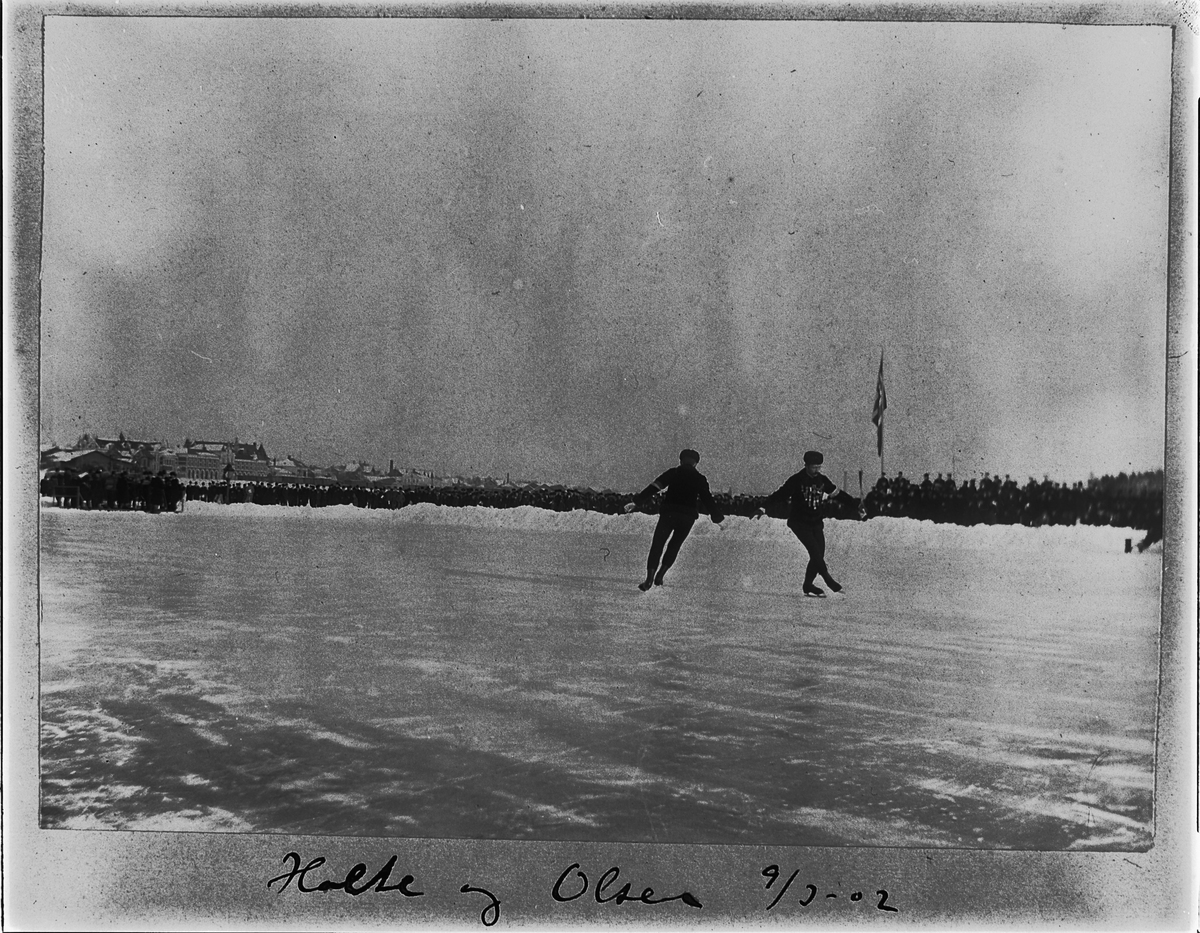 Johan Albert Steen, SKØYTELØP PÅ MJØSISEN, KUNSTLØP, OPPVISNING, OSCAR HOLTE OG PAUL OLSEN, TRIBUNE, PUBLIKUM, FLAGG, 9-3-1902, BEBYGGELSEN I STRANDGATA, HAMAR I BAKGRUNNEN.