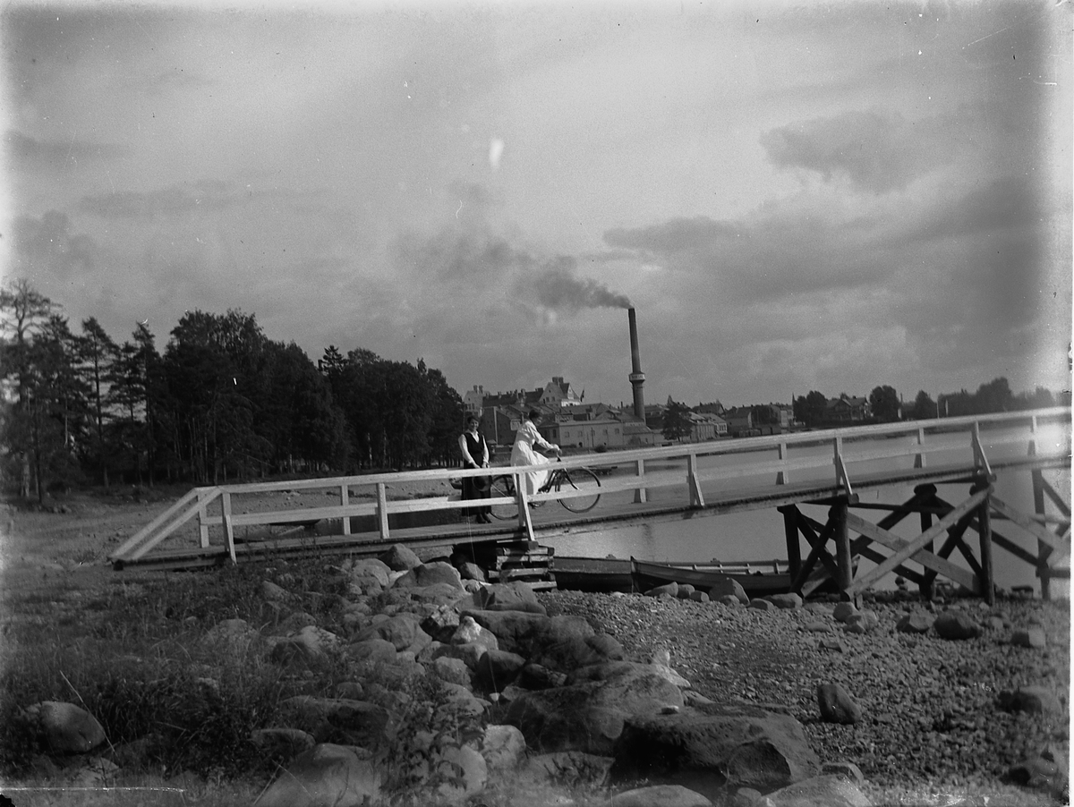 Johs. (Johannes) Johannessen, To kvinner, den ene på sykkel, ved vannkanten til Mjøsa. Brygga, gangbrua til badehuset? I bakgrunnen Hamar.