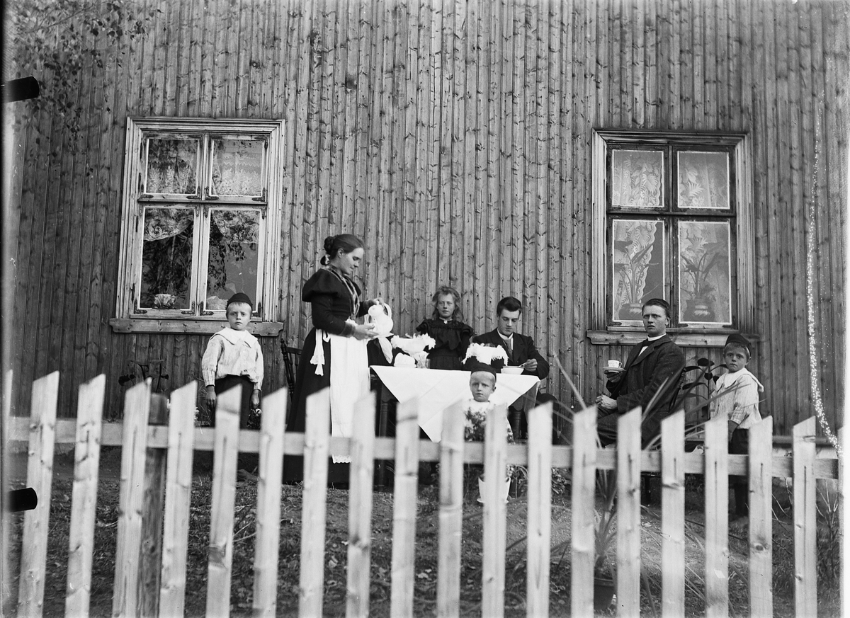 Mina Skraastad, En gruppe, familie med fire barn i en husvegg. Ei kvinne serverer kaffe.