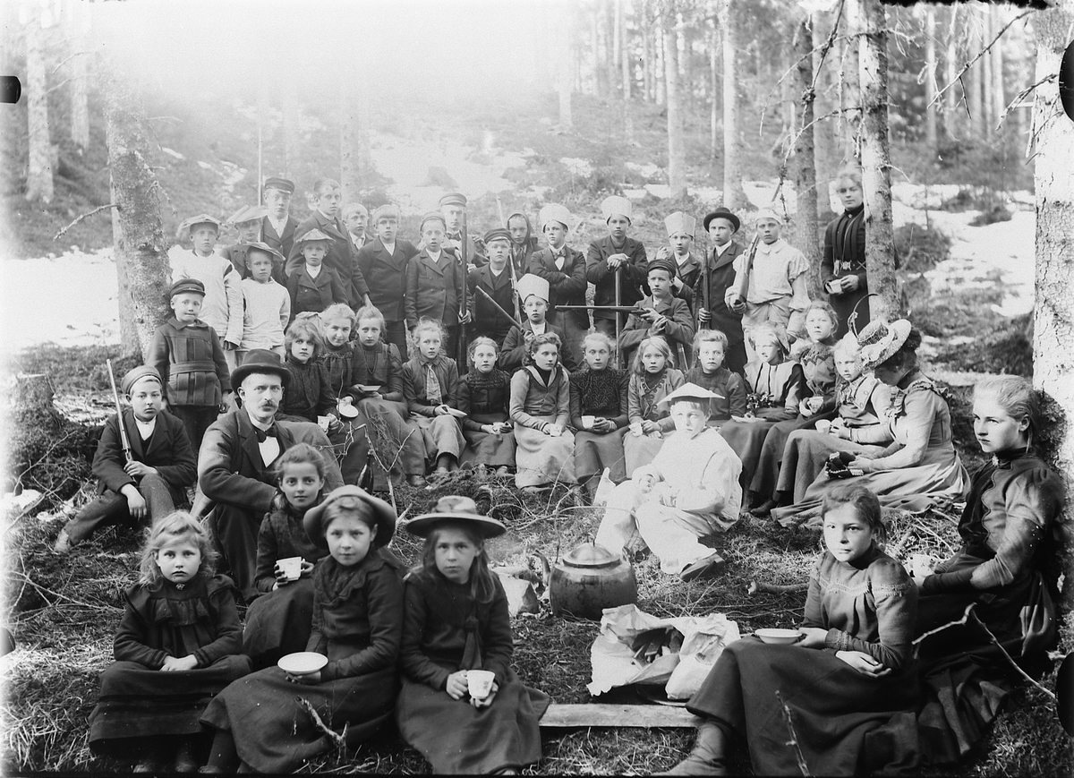 Mina Skraastad, Stor gruppe gutter og jenter, med en mann og ei kvinne, i skogen. Drikker kaffe, spiser niste. De bakerste har våpen og "hatter". Se HHB-3130.