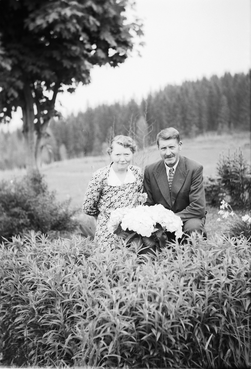 Håkon Prestkværn, Ukjent kvinne og mann med blomster i en hage, jorde og skog i bakgrunnen.